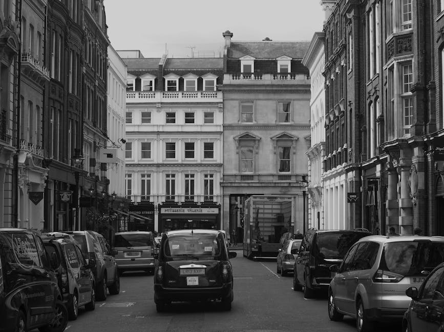 A street view in a city neighbourhood featuring a large, white, multi-story residential building with classic architectural details, including balconies with wrought iron railings and decorative windo