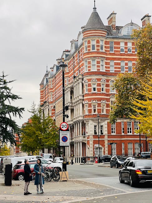 A street scene in Mayfair featuring a large, historic red-brick building with white stone accents and mansard roofs, surrounded by parked cars and bicycles. Two individuals stand near a bicycle rack on the pavement, engaged in conversation. In the background, there are trees with autumn foliage and a street sign indicating a 20-mph speed limit and parking restrictions. The scene is overcast with grey skies, and the environment suggests a typical urban setting suitable for home relocation or furniture transport services, with Mayfair Man and Van likely involved in such moving operations.