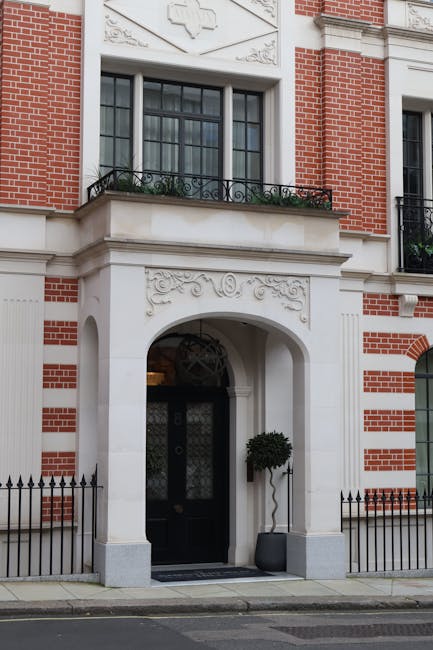 The exterior of a red-brick and white stone townhouse featuring an arched entranceway with decorative carvings above the doorway. The black front door is adorned with a glass panel and is flanked by a potted topiary tree on the right side. Above the entrance, there is a large window with black framing and a small balcony with ornate black wrought iron railings, decorated with flower boxes containing greenery. The building has a low black metal fence along the sidewalk, and the pavement appears clean and well-maintained. This setting illustrates a typical London residential building ready for home relocation or furniture transport, as part of professional removals service offered by Mayfair Man and Van, supporting efficient packing and loading processes.