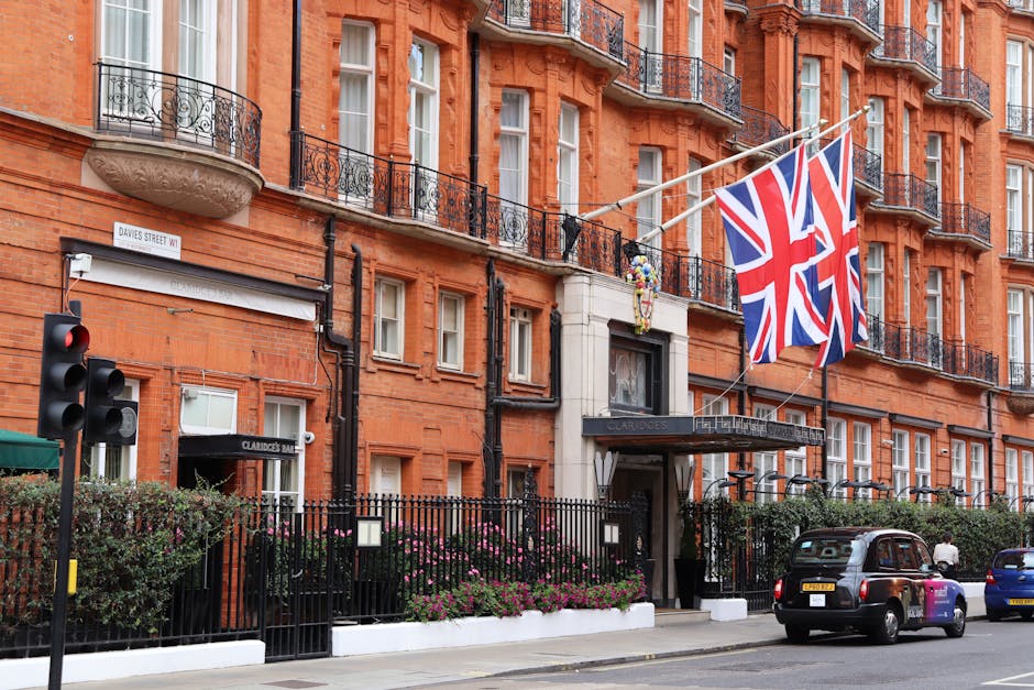 The exterior of a red-brick building with multiple sash windows and ornate black wrought iron balconies, situated on a city street with a traffic light in the foreground. Two British flags are prominently displayed above the entrance, which is framed by white stone and a black awning bearing the name 'Claridges.' In front of the building, there is a black metal fence with shrubbery and flowering plants behind it. Parked on the street are a black taxi and a blue car, alongside pedestrians walking on the sidewalk. Visible equipment includes a trolley, and a person is seen carrying or loading furniture or boxes towards the building, indicating a home relocation or furniture transport process. This scene illustrates the logistics involved in house removals, with items being moved in or out of the property, possibly connected to professional moving services provided by Mayfair Man and Van.