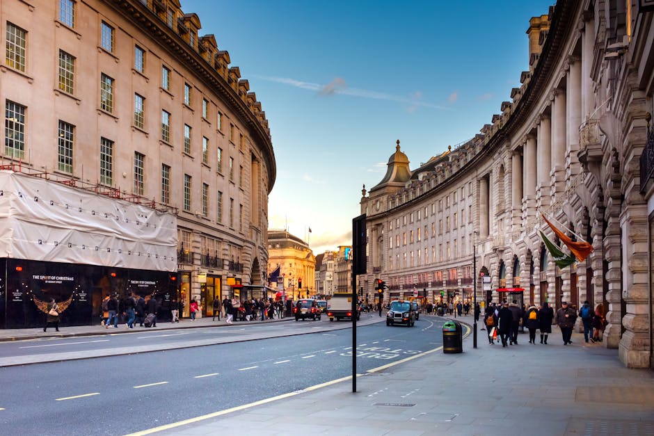 A city street scene during daytime with a curved row of historic, multi-storey buildings featuring ornate stone facades and large windows. The buildings are illuminated by natural light, with some areas under partial shade. Pedestrians are walking along the wide sidewalk, some waiting at crossing points, while others are engaged in conversations or carrying shopping bags. Several vehicles, including black cabs, taxis, and a white van, are traveling along the road, which has multiple lanes marked with white dashed lines and tram tracks embedded in the pavement. A shopping store with a large display window is visible on the left side, partially covered by a white protective sheet, indicating possible renovations. A street sign is positioned centrally, and trash bins are placed along the sidewalk. The scene depicts typical urban activity related to house removals and packing logistics in central London, with potential for moving services such as furniture transport and home relocation, as offered by Mayfair Man and Van.