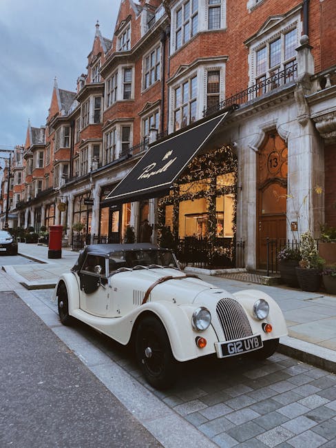 A vintage cream-colored convertible car with black accents and a black soft top is parked on a residential street in front of a row of red-brick terraced houses with white window frames and ornate stone detailing. The car’s registration plate reads 'G12 UVB.' The street is paved with grey bricks, and a red postbox is visible further down the pavement. In the background, there is a property with a black awning that has the word 'Hotel' written on it, partially obscured by trees and decorative lighting. The scene is captured during the late afternoon or early evening, with soft natural light illuminating the surroundings. This image depicts a typical urban setting where house removals or furniture transport might take place, with furniture or items possibly being loaded or unloaded nearby as part of a home relocation process, supported by professional removals services such as those offered by Mayfair Man and Van.