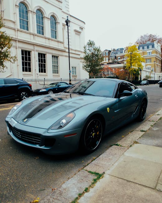 A dark grey Ferrari sports car with a sleek, aerodynamic design is parked on the street adjacent to a sidewalk in front of a large, white, classical-style building with tall arched windows and a black metal fence. The car's tinted windows and distinctive yellow Ferrari emblem are visible, and it features black alloy wheels with yellow brake calipers. In the background, several other vehicles are parked along the street, and a row of trees with mixed green and yellow foliage line the sidewalk, indicating an autumn setting. The scene appears to be during daytime with diffused natural light, and the area suggests an affluent neighbourhood suitable for professional house moves where Mayfair Man and Van might provide removals and furniture transport services, including packing and loading processes.