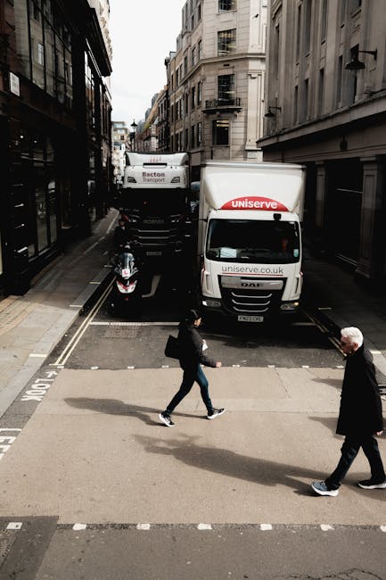 A street scene in an urban area with tall, stone-faced buildings lining both sides of the street. In the foreground, two pedestrians are walking across a zebra crossing; one person is wearing a black jacket and jeans, carrying a backpack, while an older man with white hair is dressed in dark clothing. Behind them, a white moving van with the logo and website of uniserve.co.uk is parked against the curb, partially blocking the street. Further back, a larger truck from Bexton Transport is visible, situated behind the van, both positioned across the narrow road that is used for vehicle loading and unloading. The scene suggests a home relocation or furniture transport process in progress, consistent with professional removals services provided by Mayfair Man and Van. The ambient lighting indicates daytime, with overcast skies casting a neutral light on the scene.
