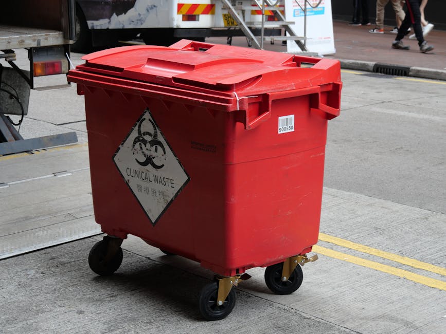A red wheeled clinical waste bin with a hinged lid, positioned on a concrete pavement near a vehicle used for house removals. The bin features a biohazard label with the words 'Clinical Waste' and a triangular hazard symbol. It is situated at the edge of a property, with a yellow line marking the boundary between the pavement and the road. In the background, a staircase leading to an elevated area and a group of people walking on the pavement are partially visible. The scene suggests waste collection as part of a home relocation or packing process, with the bin prepared for disposal of medical or hazardous materials during house removals, with Mayfair Man and Van providing comprehensive moving services.