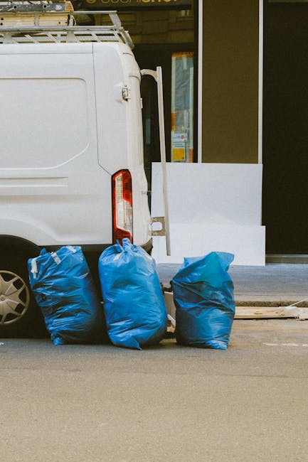 A white commercial van parked on a street outside a building with a glass entrance door, partially visible. Four large blue plastic bags filled with items are placed on the pavement next to the rear o