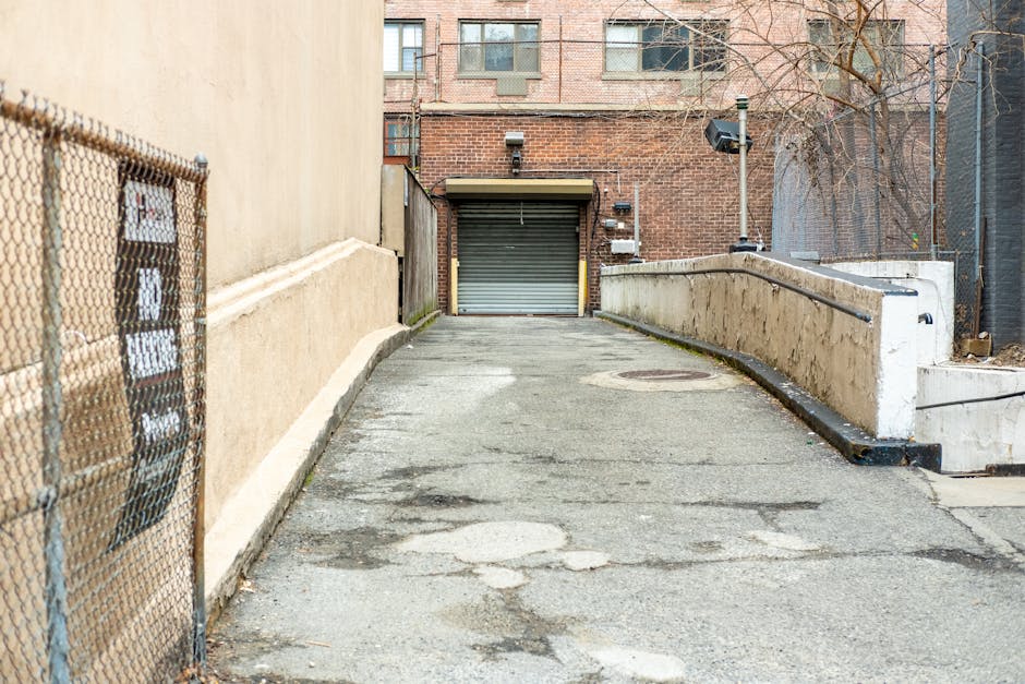 A concrete driveway leading up to a garage with a roll-up metal door at the end of an alleyway, flanked by a beige building on the left and a black metal fence on the right. The driveway is slightly uneven with visible patches and cracks, and it slopes upward towards the garage entrance. To the right, there are a white concrete ramp with a metal handrail, leading to an internal doorway, which appears to be used for home relocation or furniture transport access, typical of move-in or move-out logistics. In the background, a brick building with multiple windows and a metal exhaust vent is visible. A security camera is mounted on a pole near the ramp, and there is minimal natural light, suggesting daytime. The scene depicts an outdoor loading area used by professional removal services, such as Mayfair Man and Van, for executing house removals and transportation of household items.