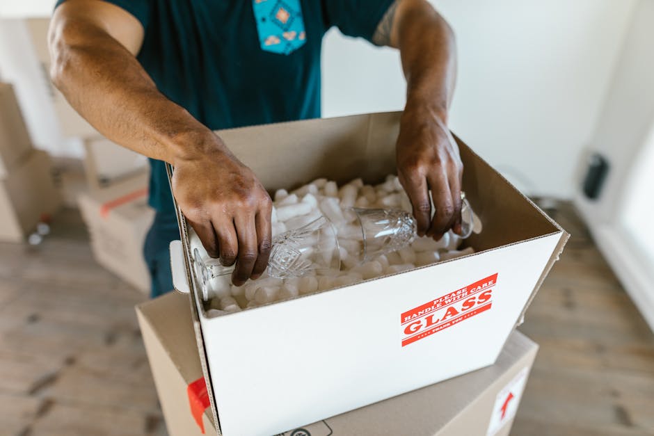 A person wearing a blue shirt is seen packing or unpacking a cardboard box labeled 'Glass' with red printing, filled with protective white foam packing peanuts. The individual is holding a clear glass bottle, positioning it carefully within the box for a home relocation or furniture transport process. The setting appears to be inside a residential property, with wooden flooring visible and soft natural lighting illuminating the scene. The box is placed on top of a larger, partially visible moving box or container. This activity reflects the packing and moving phase involved in a professional removals service, such as those provided by Mayfair Man and Van, focused on handling delicate valuables like glassware safely during the loading process.