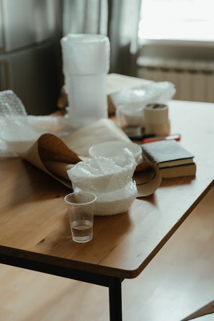 A wooden table inside a home with several packing materials and moving supplies arranged on its surface. There are large rolls of bubble wrap, crumpled bubble wrap ready for cushioning, a transparent plastic cup, and a few cardboard boxes. In the background, a window lets in natural light, and a radiator is visible beneath it. The scene depicts the packing process typically involved in house removals, with the supplies prepared for safely transporting household items. The setup suggests an ongoing packing or moving activity facilitated by Mayfair Man and Van, a professional removals service specialising in home relocation and furniture transport in Mayfair.