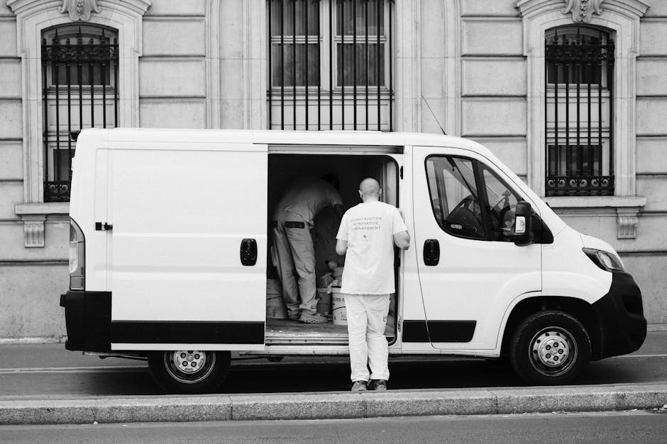 A white panel van is parked on the street in front of a historic stone building with tall, narrow windows secured by black iron bars. Three movers, dressed in light clothing, are actively engaged in t