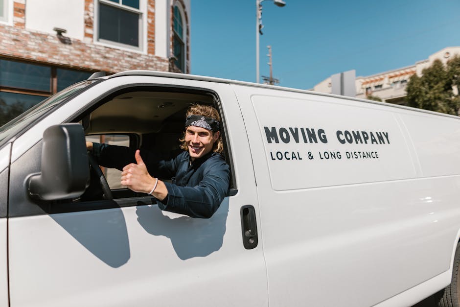 A white panel van is parked on the street in front of a historic stone building with tall, narrow windows secured by black iron bars. Three movers, dressed in light clothing, are actively engaged in t