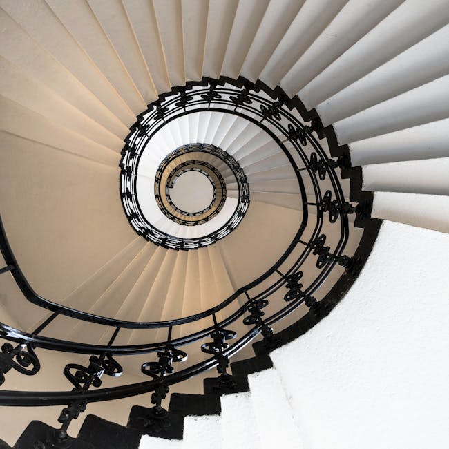 A top-down view of a spiral staircase inside a residential property, featuring white steps with black edges and a black wrought iron balustrade that follows the curvature of the staircase. The staircase descends in a tight spiral towards the central opening, with soft, natural lighting illuminating the white walls and steps. The image captures the elegant design and structural complexity of the staircase, illustrating a space where careful furniture transport and home relocation processes might occur during a move managed by Mayfair Man and Van, a professional removals service. The setting emphasizes the importance of precise packing and careful navigation in confined staircase environments typical of Mayfair's historic buildings, highlighting the need for expert handling during furniture transport and home packing efforts.