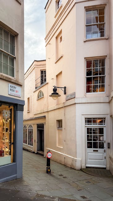 A narrow street in Mayfair with cream-colored Georgian-style buildings featuring multiple sash windows and decorative architectural details. In the foreground, there is a small shop with large display windows, and a black outdoor lamp mounted on the building wall. The street is paved with grey cobblestones, and a red and white bollard is positioned on the pavement. The image appears to be taken during daylight with partly cloudy skies. This setting depicts a typical scene for home relocation or furniture transport in a historic urban area, suitable for professional removals services like those provided by Mayfair Man and Van, who specialise in house removals and logistical support for moving within tight city streets.