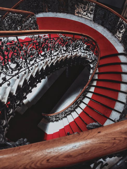 A top-down view of a spiral staircase inside a residential property, featuring white steps with black edges and a black wrought iron balustrade that follows the curvature of the staircase. The staircase descends in a tight spiral towards the central opening, with soft, natural lighting illuminating the white walls and steps. The image captures the elegant design and structural complexity of the staircase, illustrating a space where careful furniture transport and home relocation processes might occur during a move managed by Mayfair Man and Van, a professional removals service. The setting emphasizes the importance of precise packing and careful navigation in confined staircase environments typical of Mayfair's historic buildings, highlighting the need for expert handling during furniture transport and home packing efforts.