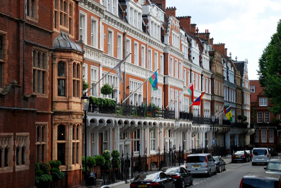 A row of historic red-brick terraced buildings in Mayfair, London, featuring ornate white window frames, decorative architectural detailing, and small balconies with black wrought iron railings. Several flags are displayed on flagpoles attached to the building facades, including a blue flag with a white emblem, a red and blue flag with a gold emblem, and others. The street in front includes parked cars of various sizes and colours, and the pavement is lined with lush green trees. The scene is captured during daylight with overcast sky, highlighting the exterior of the buildings and their classic London townhouse appearance, relevant to house removals and relocation services provided by Mayfair Man and Van.