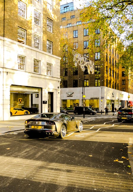 A street scene in Mayfair showing the exterior of multi-storey buildings with large windows, some with reflective glass panels, and decorative street lighting hanging above the road. The pavement features trees with green and yellow leaves, indicating an autumn setting. parked directly in front of the buildings are several luxury vehicles, including a distinctive grey sports car with a sleek, aerodynamic design and visible rear lights, and a yellow sports car inside a vehicle showroom with large glass windows. The street has marked pedestrian crossings, and a few pedestrians are walking along the sidewalk. The scene captures the urban environment typical of Mayfair, with a focus on high-end transport and architecture, relevant for house removals and furniture transport services provided by Mayfair Man and Van.
