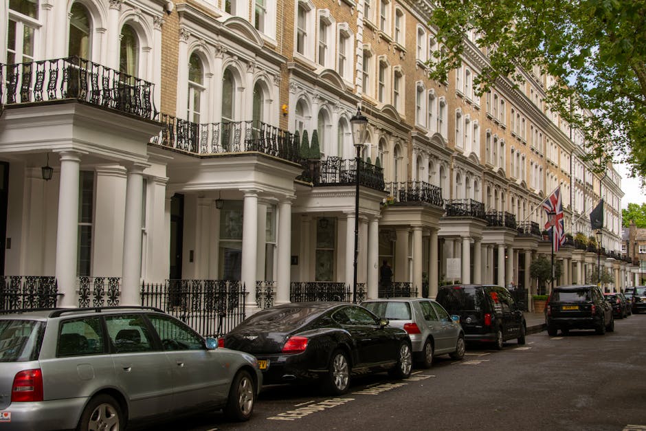 A row of historic red-brick terraced buildings in Mayfair, London, featuring ornate white window frames, decorative architectural detailing, and small balconies with black wrought iron railings. Several flags are displayed on flagpoles attached to the building facades, including a blue flag with a white emblem, a red and blue flag with a gold emblem, and others. The street in front includes parked cars of various sizes and colours, and the pavement is lined with lush green trees. The scene is captured during daylight with overcast sky, highlighting the exterior of the buildings and their classic London townhouse appearance, relevant to house removals and relocation services provided by Mayfair Man and Van.